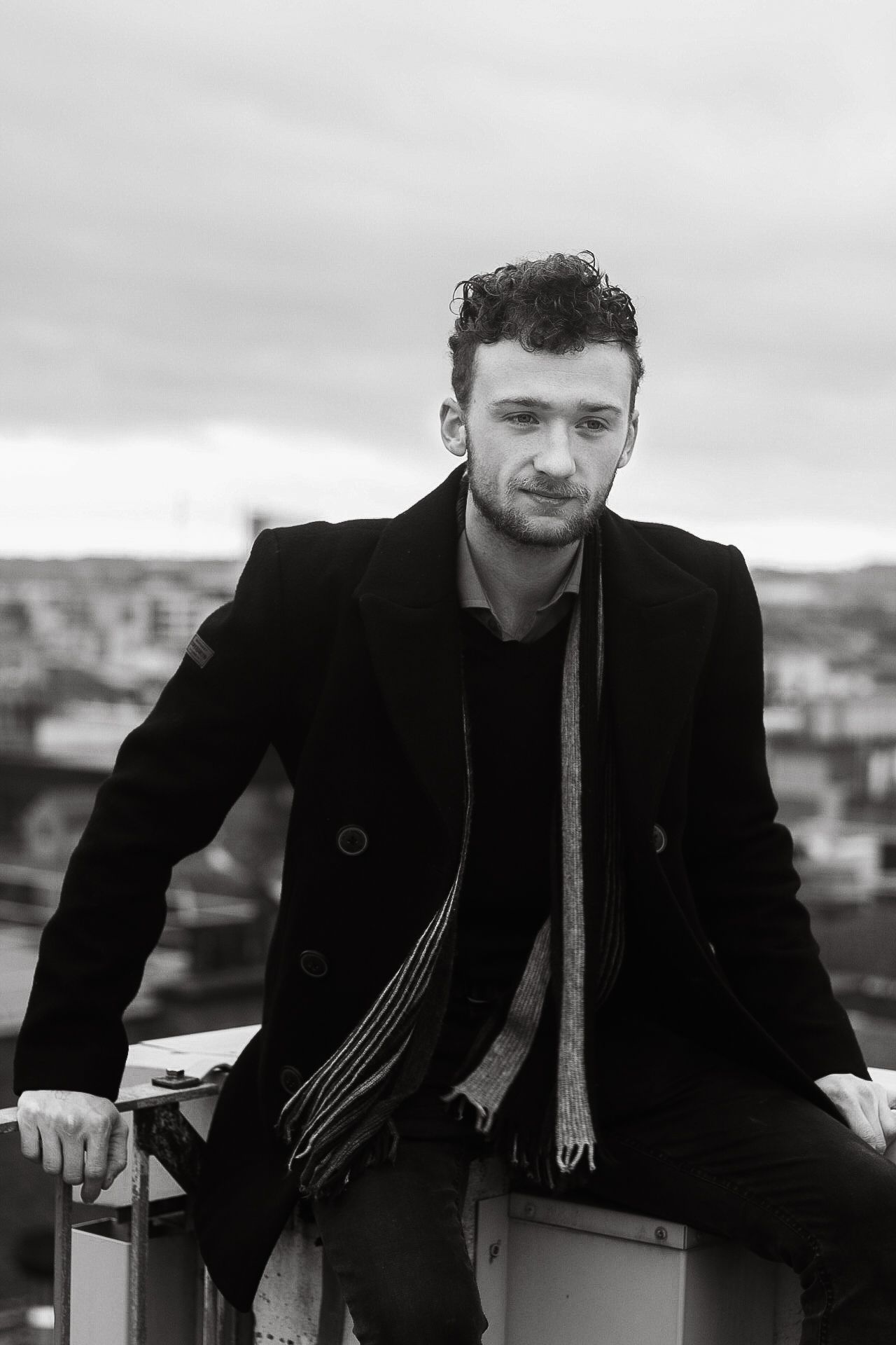 A moody black and white portrait of a person in a winter coat against a cityscape backdrop.