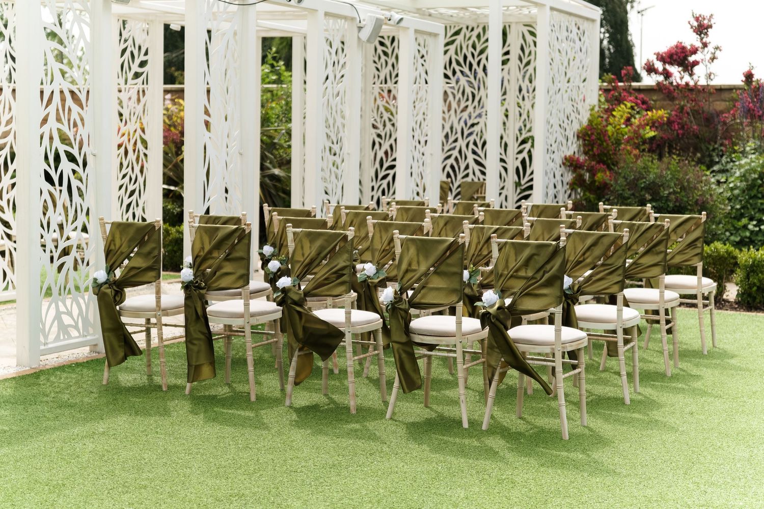 Elegant outdoor wedding ceremony chairs with olive green sashes and white floral accents on green grass near decorative white screens.
