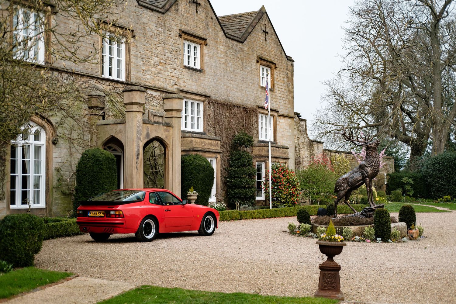 Red Porsche 944 parked on gravel driveway of elegant English country manor with bronze stag sculpture.