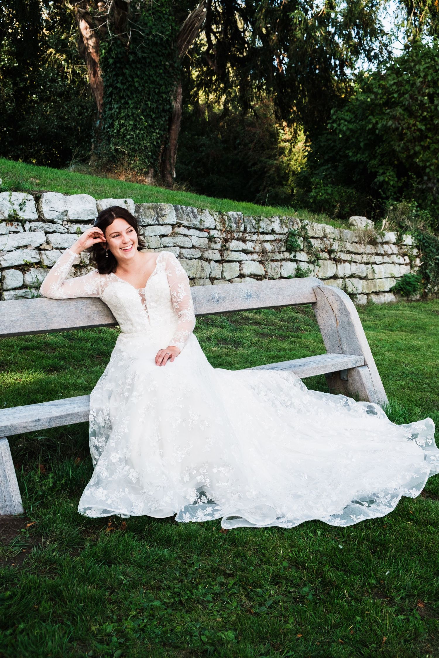 A bride in a flowing lace wedding dress reclines on a rustic stone wall bench in a garden setting.