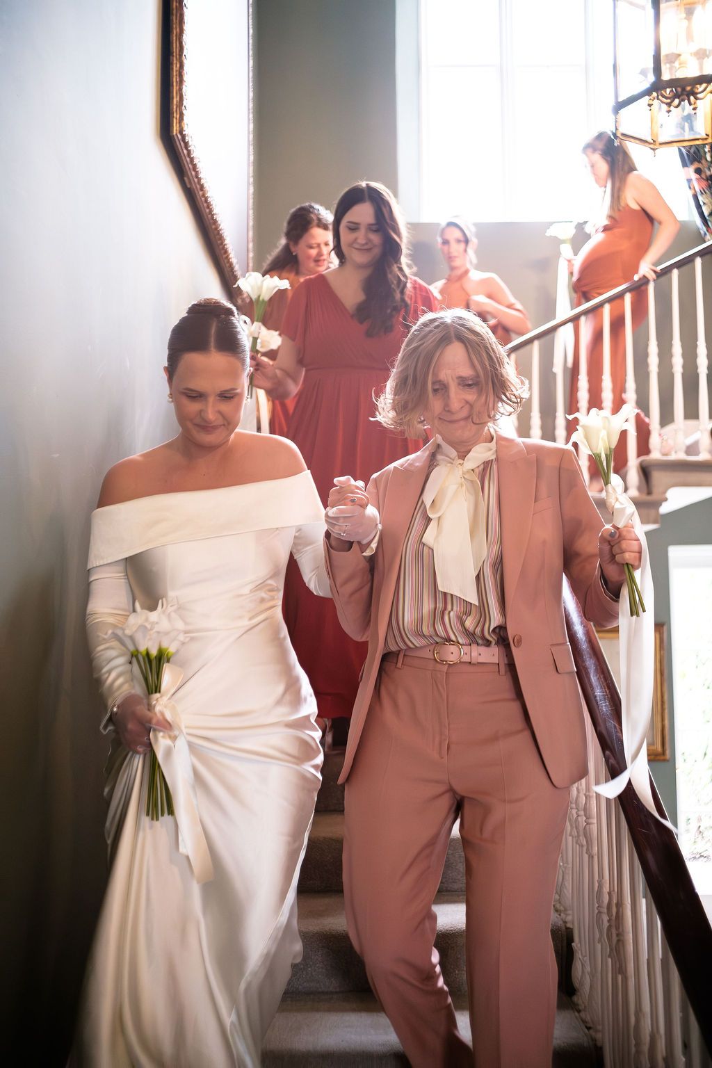 Two brides descend a staircase hand-in-hand, one in a white gown and one in a pink suit, followed by bridesmaids in red.
