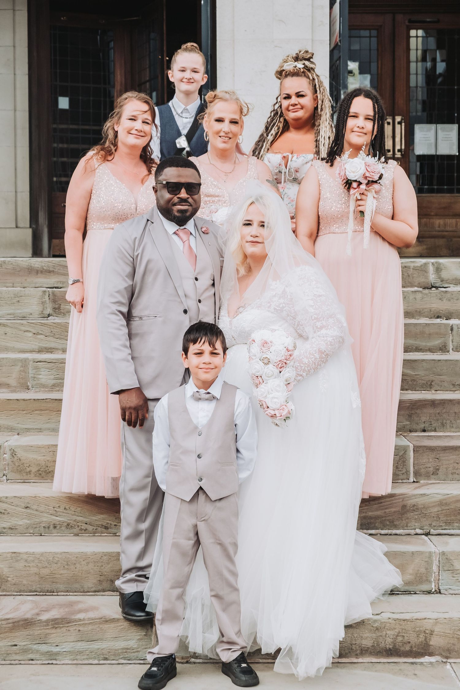 Wedding party poses on stone steps with bridesmaids in pink dresses and groom and young ring bearer in light gray suits.