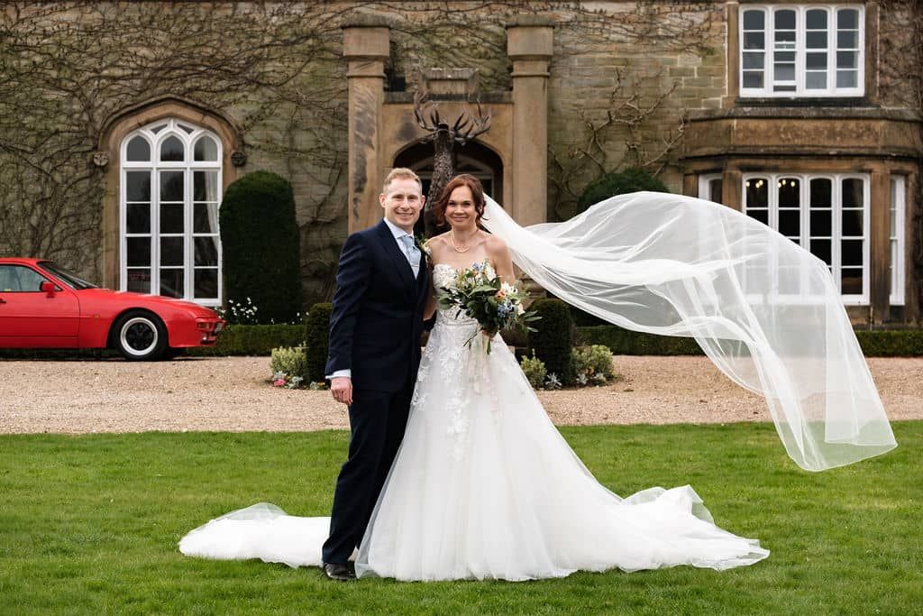 Bride and groom pose on lawn before Dunston Hall Estate, her veil blowing in wind, red car visible behind them taken by 21 Photography