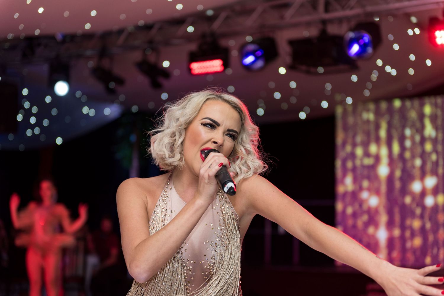 A performer in a shimmering dress gestures dramatically while singing on stage with colorful lighting shot in Leeds by 21 Photography