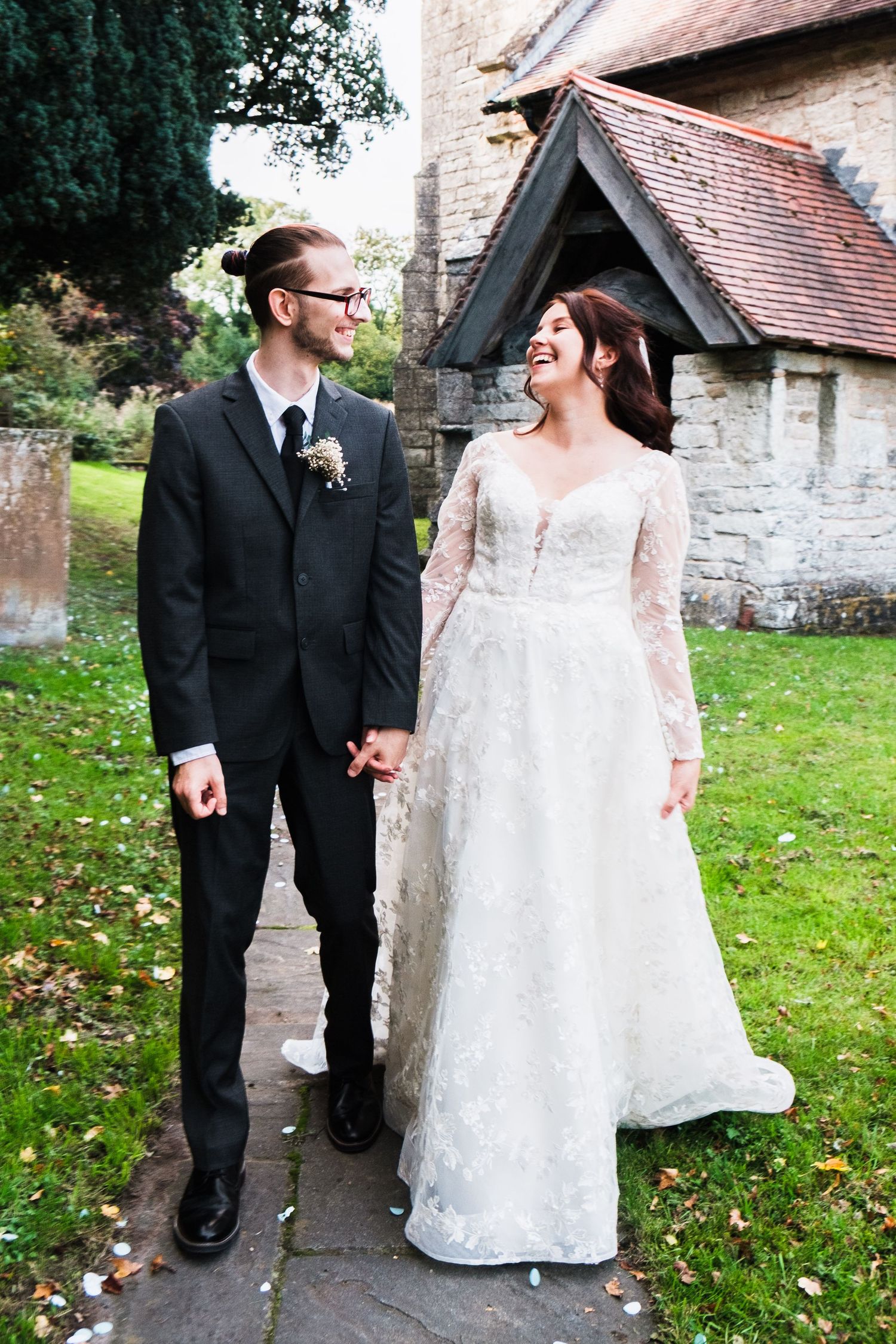 A couple in wedding attire walks together on grass near a stone church building taken by chesterfield wedding photographer 21 Photography
