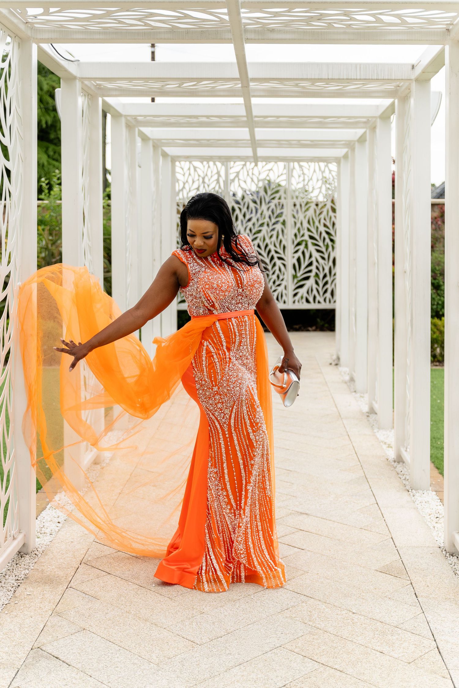 Woman in orange beaded gown with flowing cape poses gracefully in white pergola walkway. At Wildes Inn, Chesterfield taken by 21 Photography