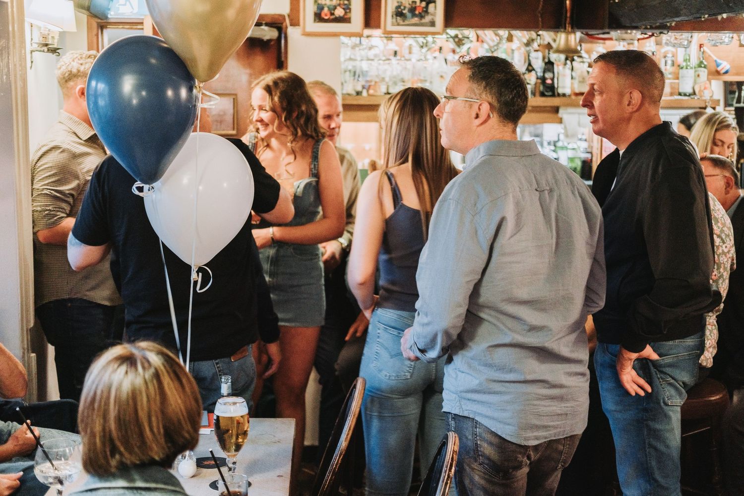 A group of people gathered at a party with decorative lights and balloons visible in the background.