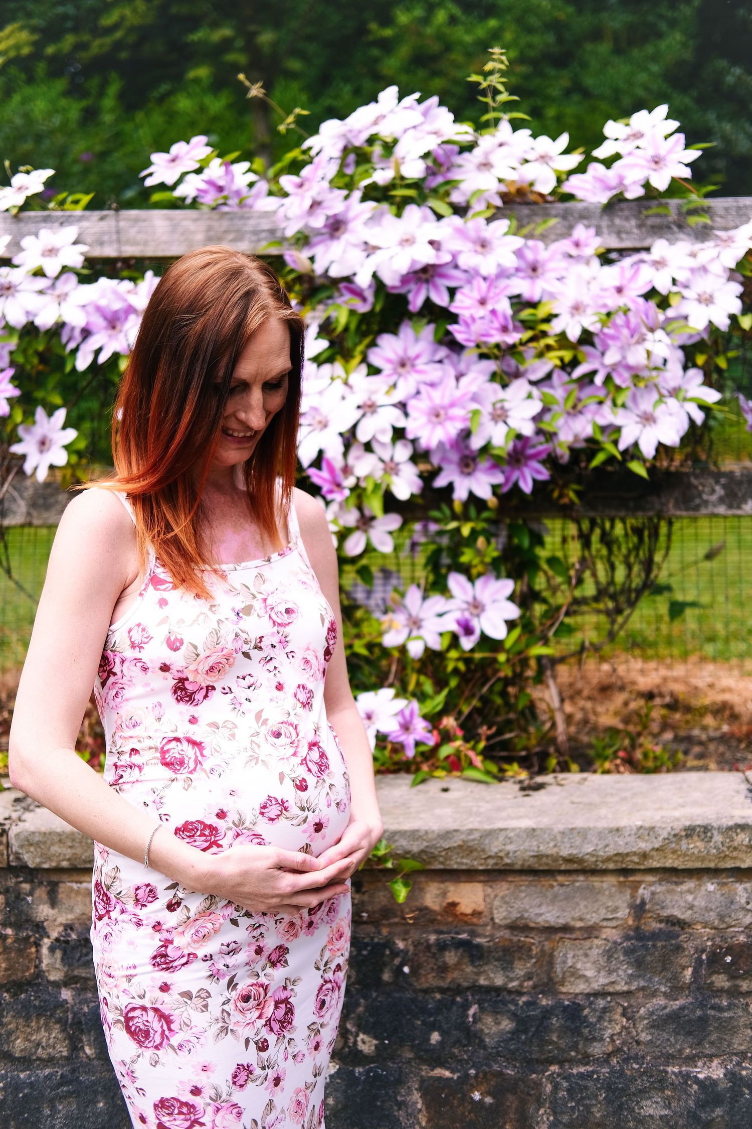 A pregnant woman in a floral dress stands by blooming clematis flowers during an outdoor photoshoot.