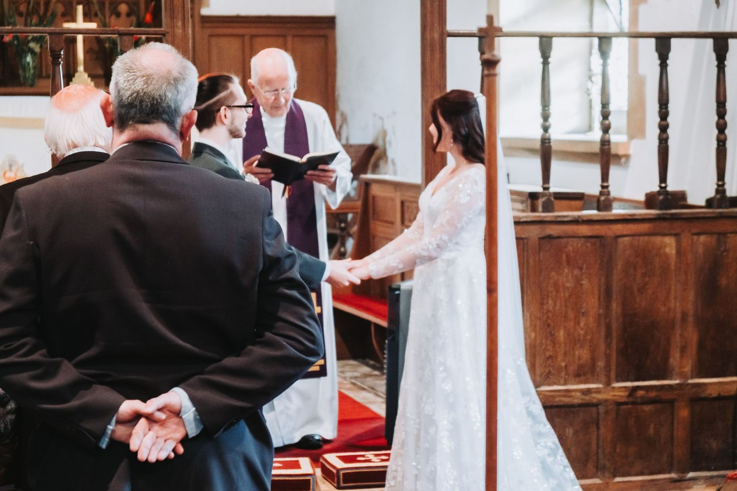 A couple holds hands during a traditional church wedding ceremony with wooden pews and stained glass windows.