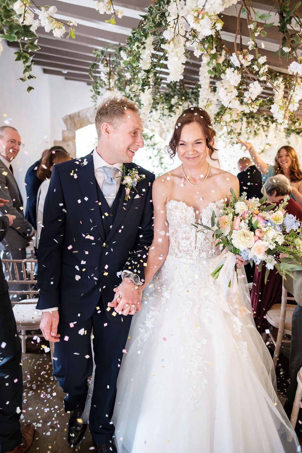Bride and groom walking through confetti under hanging blossom, both smiling