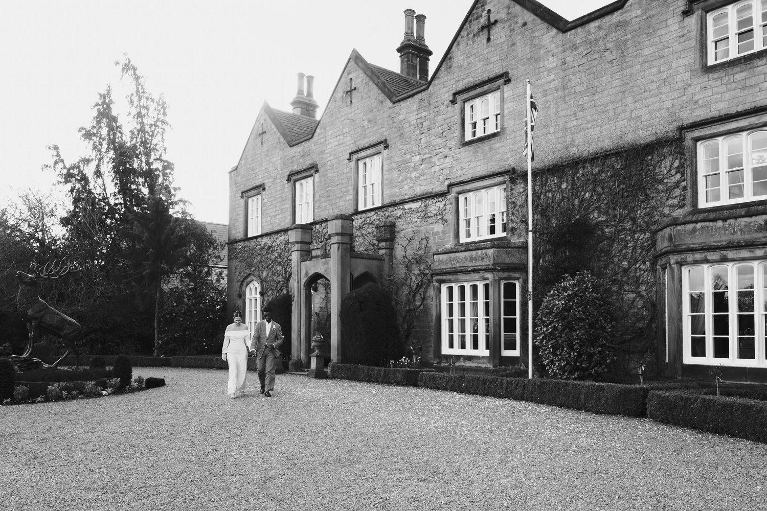 Couple walking hand in hand outside a grand stone manor house on a gravel driveway in black and white  at dunston hall estate
