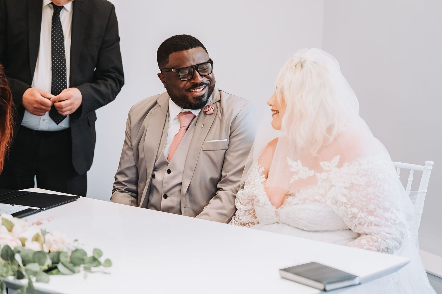 A groom in a gray suit and bride in white lace laugh together while signing wedding documents at a white table