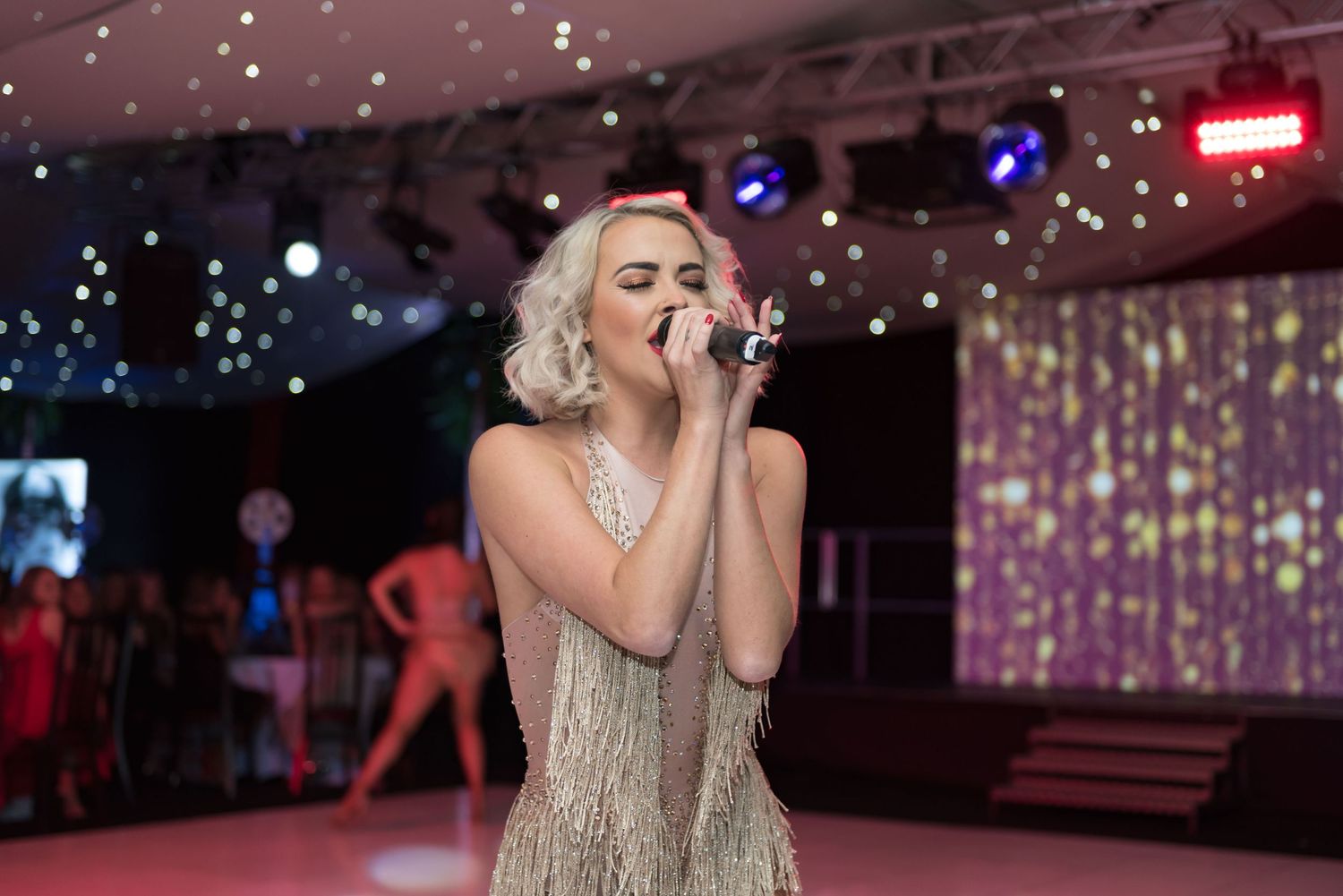 A performer in a sparkly beige dress sings passionately on stage with dramatic lighting and decorative elements shot in Leeds by 21 Photogra