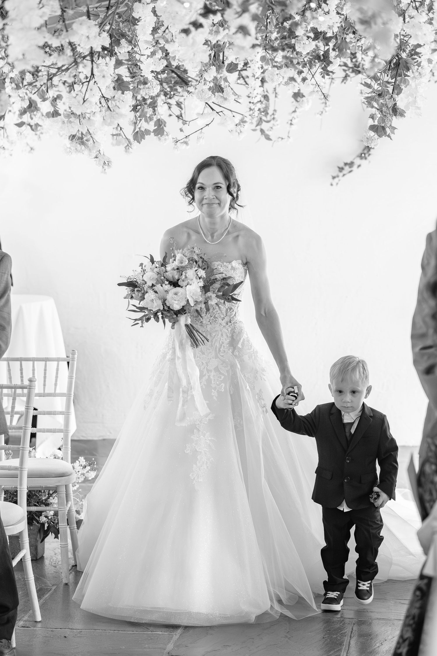 Smiling bride in lace gown walks down aisle holding bouquet and young boy's hand at wedding.