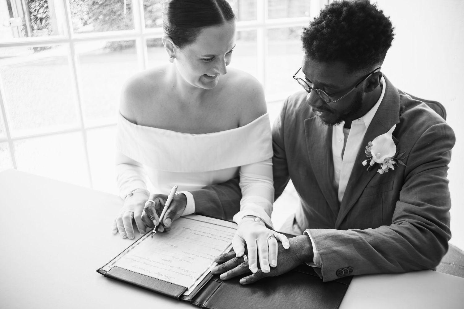 Bride and groom signing wedding registry documents at ceremony table in black and white photo.