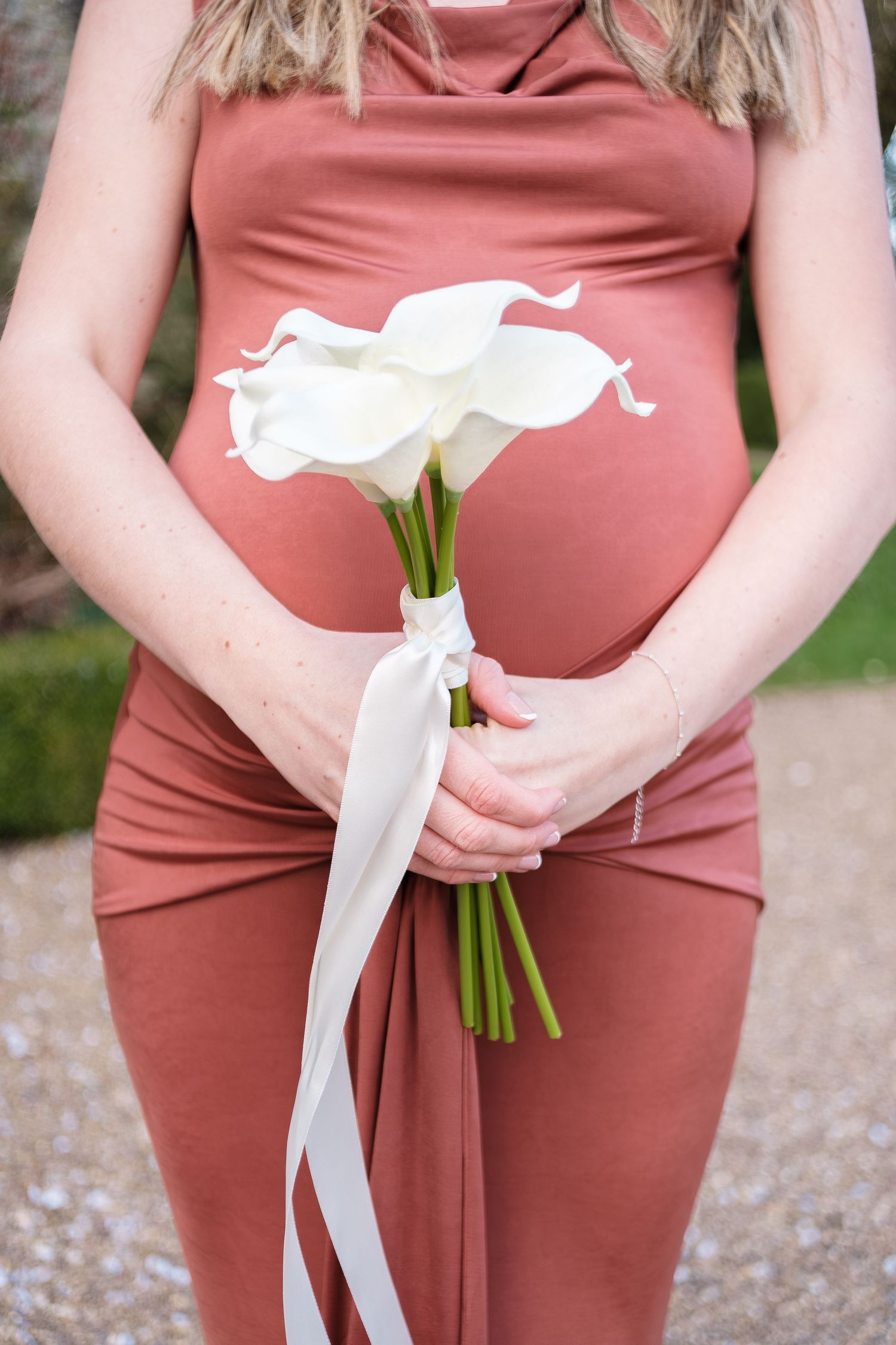 Pregnant woman in dusty rose dress holding white calla lily bouquet with ribbon, showcasing baby bump outdoors.