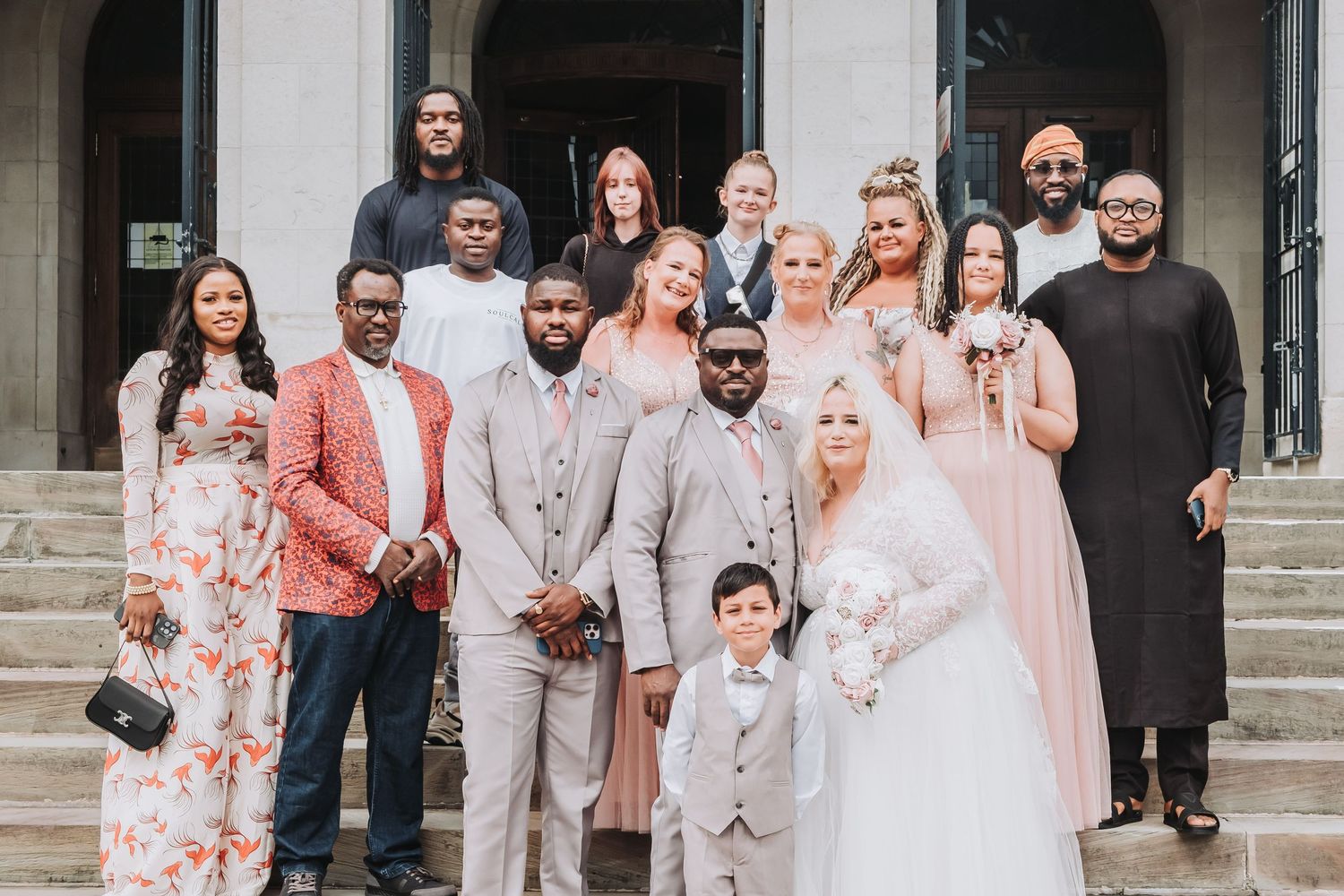 A festive wedding party in pastel and floral attire poses together on stone steps in front of a building.
