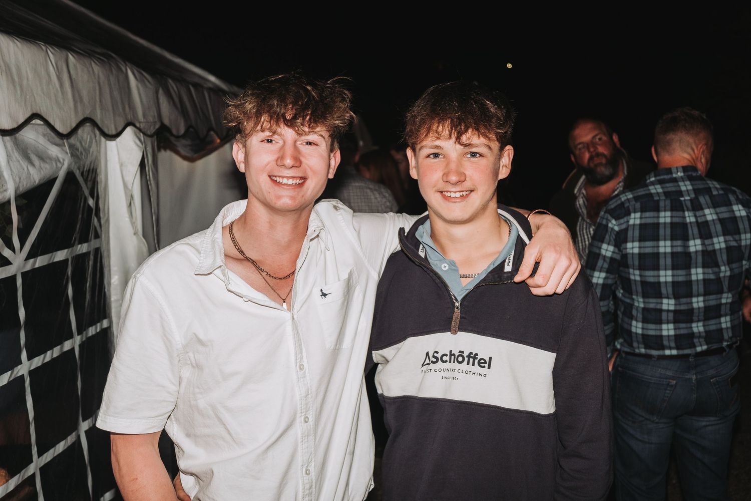 Two friends posing together at a nighttime event under a tent.