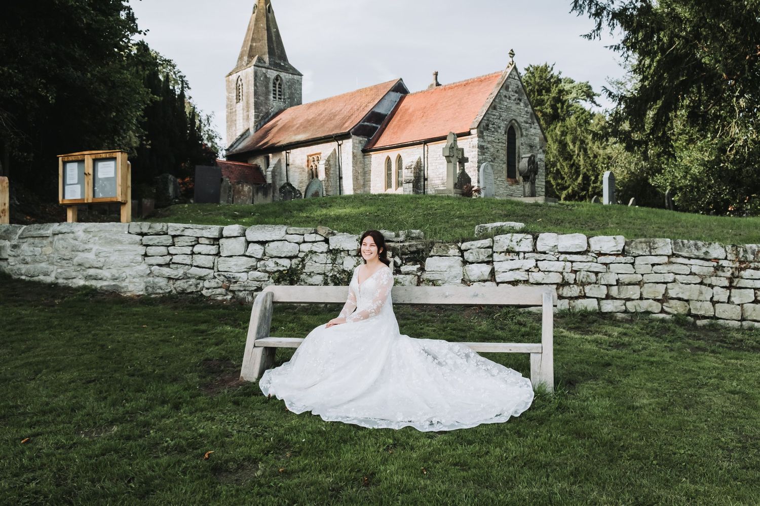 A bride in a white wedding gown poses gracefully on a stone wall surrounded by lush green gardens.