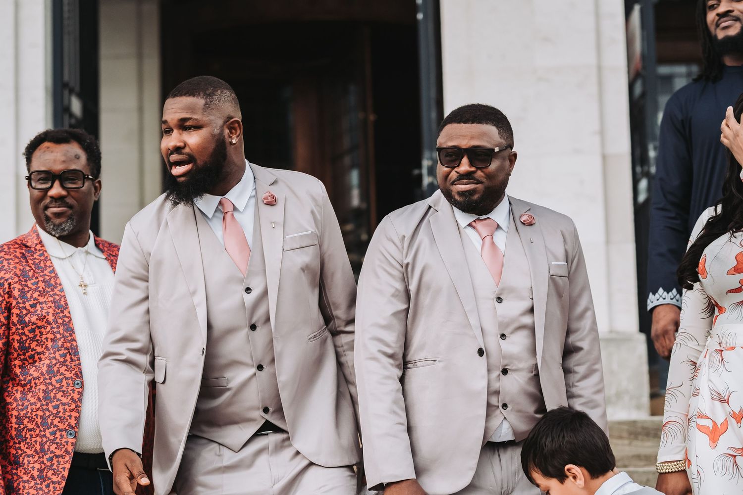 Wedding party members in coordinated grey suits and coral accessories pose on stone steps.