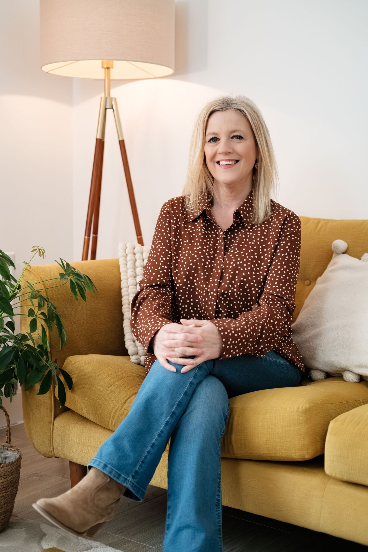 Smiling blonde woman in brown polka dot blouse and jeans sitting on yellow sofa in modern living room.