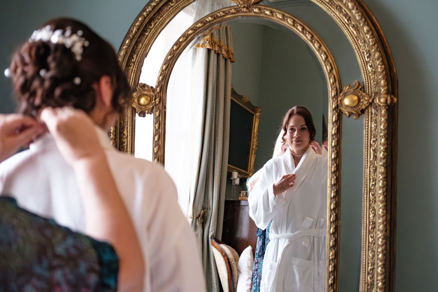 Bride in white robe adjusts earring before ornate gold mirror in elegant room.