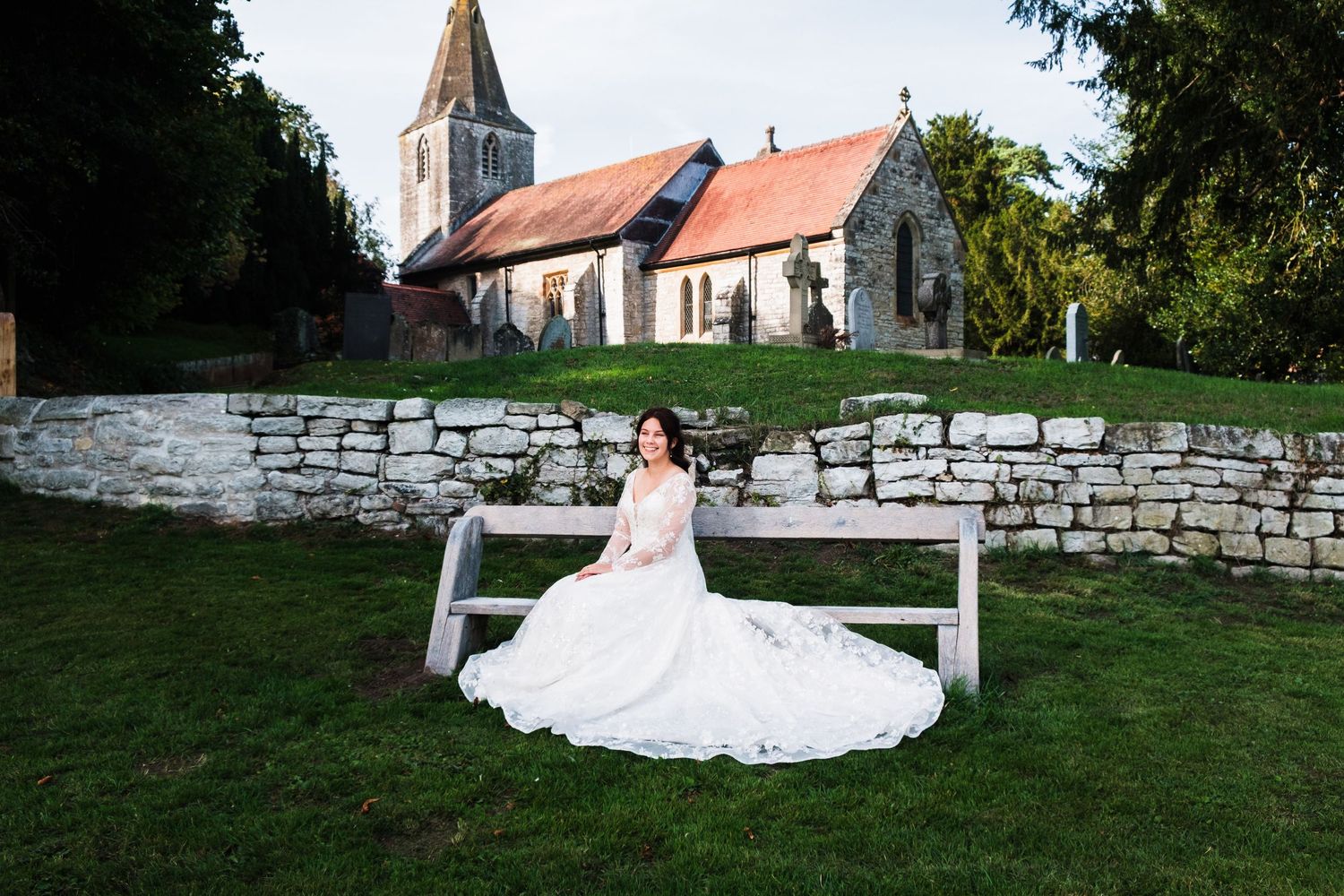 A white wedding dress flows across a wooden bench on church grounds with a stone wall and steeple in the background.