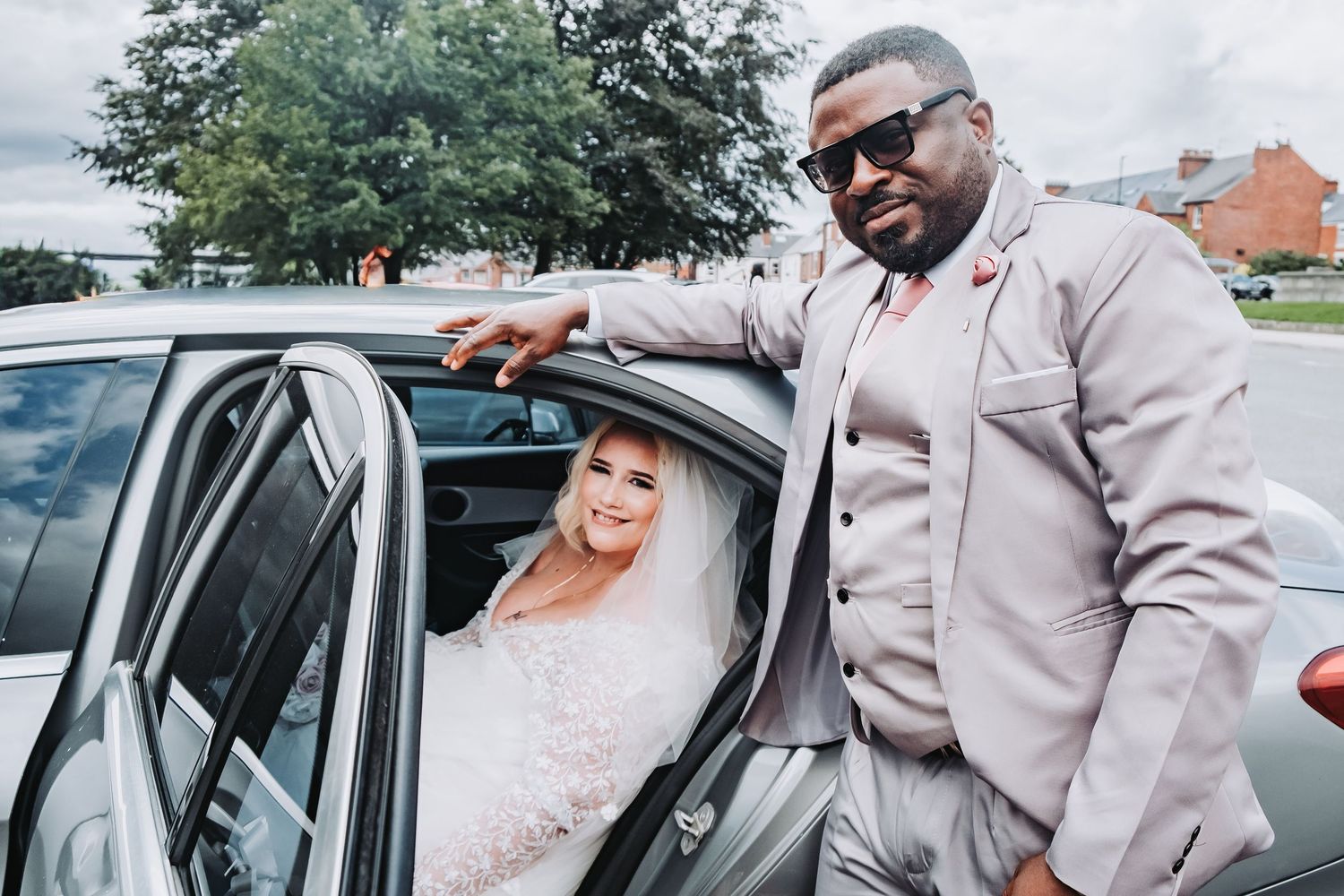 A wedding couple shares intimate moments by their car, with the bride wearing a pink dress and groom in beige suit.