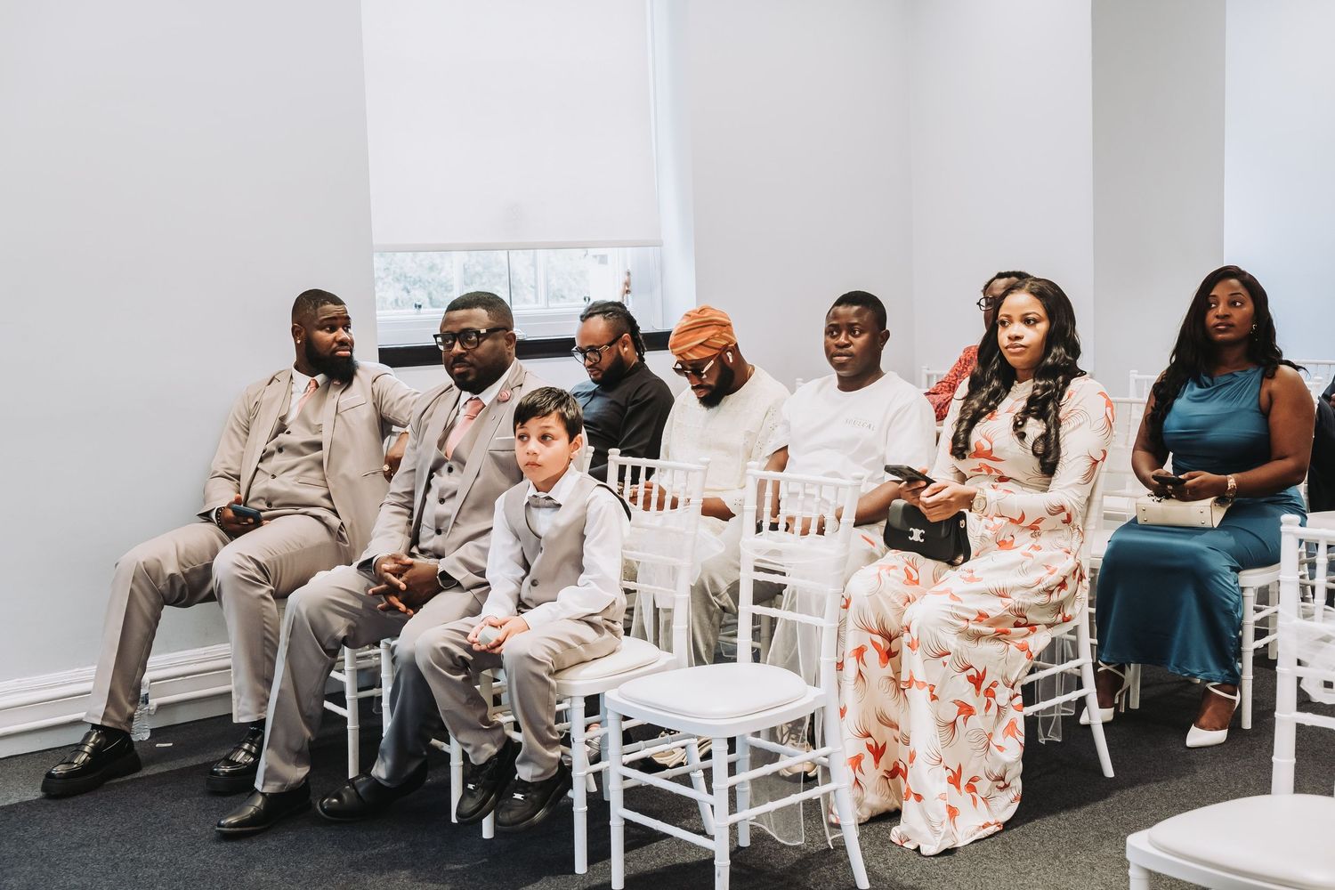Guests seated in white Chiavari chairs at a formal event wearing stylish attire in neutral and floral colors.