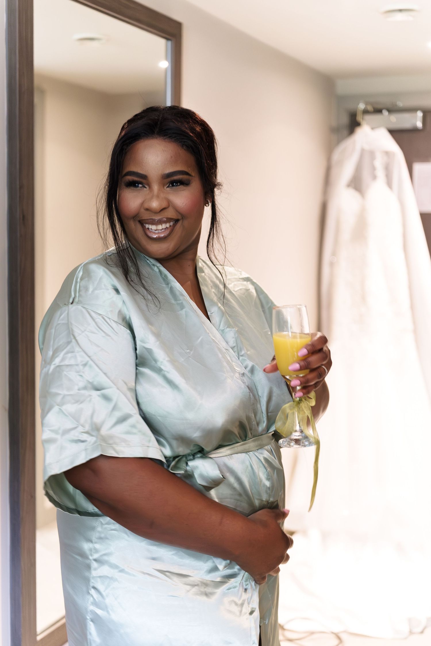 Smiling bride in mint satin robe holding champagne glass wedding dress visible in background. at Wildes Inn taken by 21 Photography