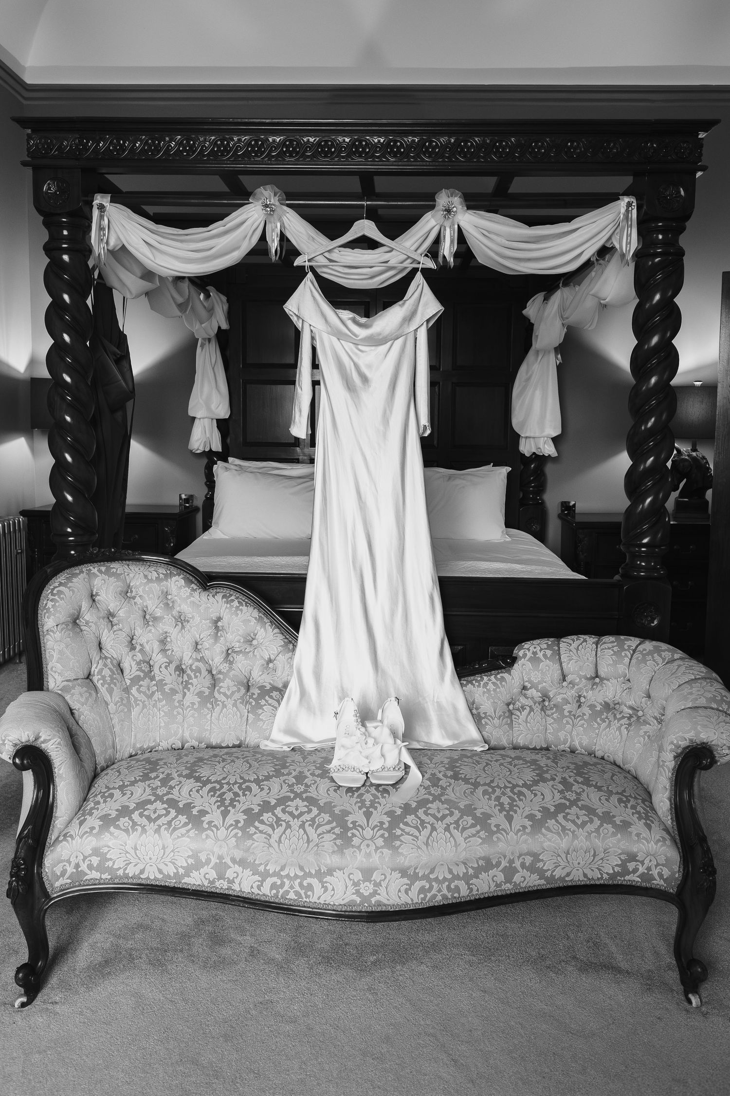 Black and white photo of an elegant wedding dress hanging on a four-poster bed above a vintage chaise lounge with bridal shoes  at dunston