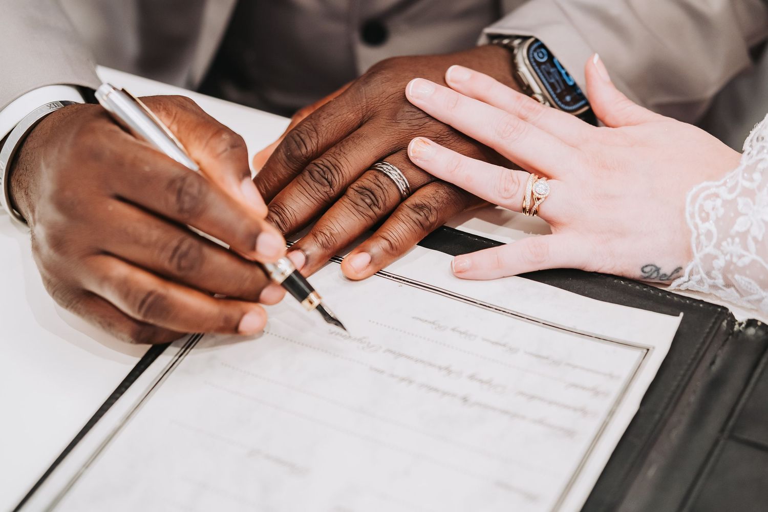 Detailed view of hands holding pens while signing marriage documents on white table.