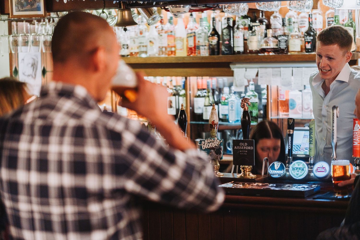 Two people enjoying drinks at a bustling bar counter with bottles and glasses visible in the background.