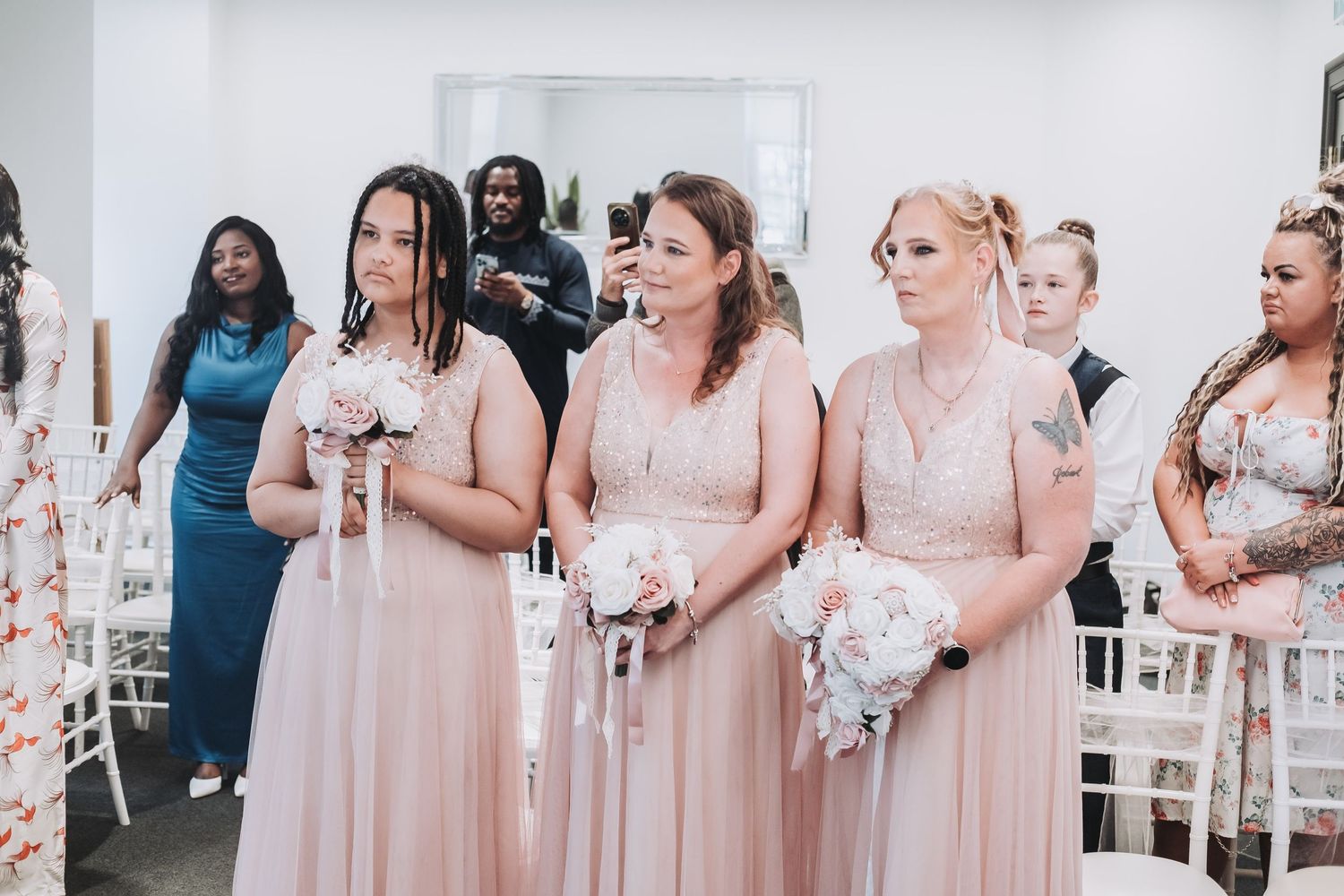 Wedding party attendees in blush pink dresses stand in a line holding bouquets at an elegant venue.