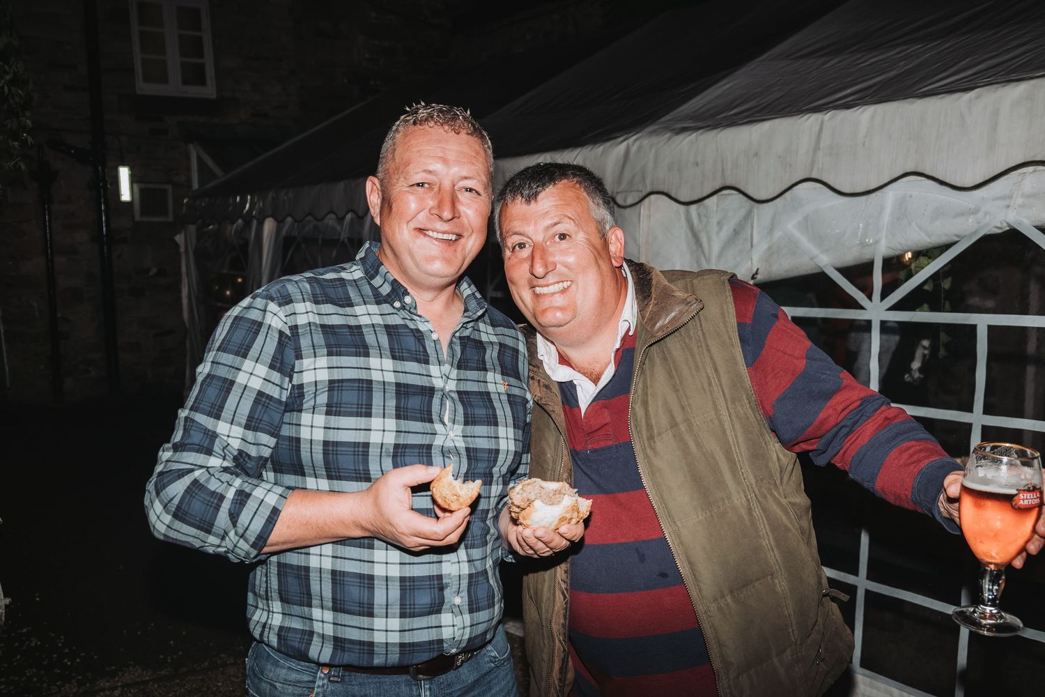 Two men smile and pose together at a nighttime gathering under a tent, one wearing plaid and one in a striped sweater.