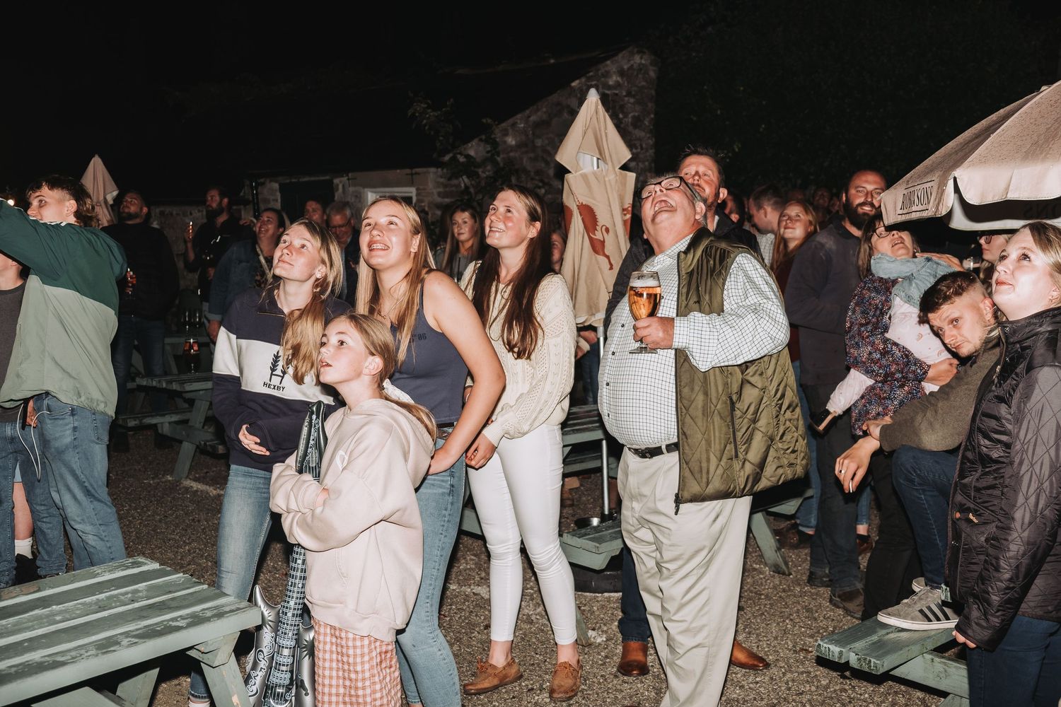 Nighttime outdoor gathering with people holding drinks and standing around picnic tables at a social event.
