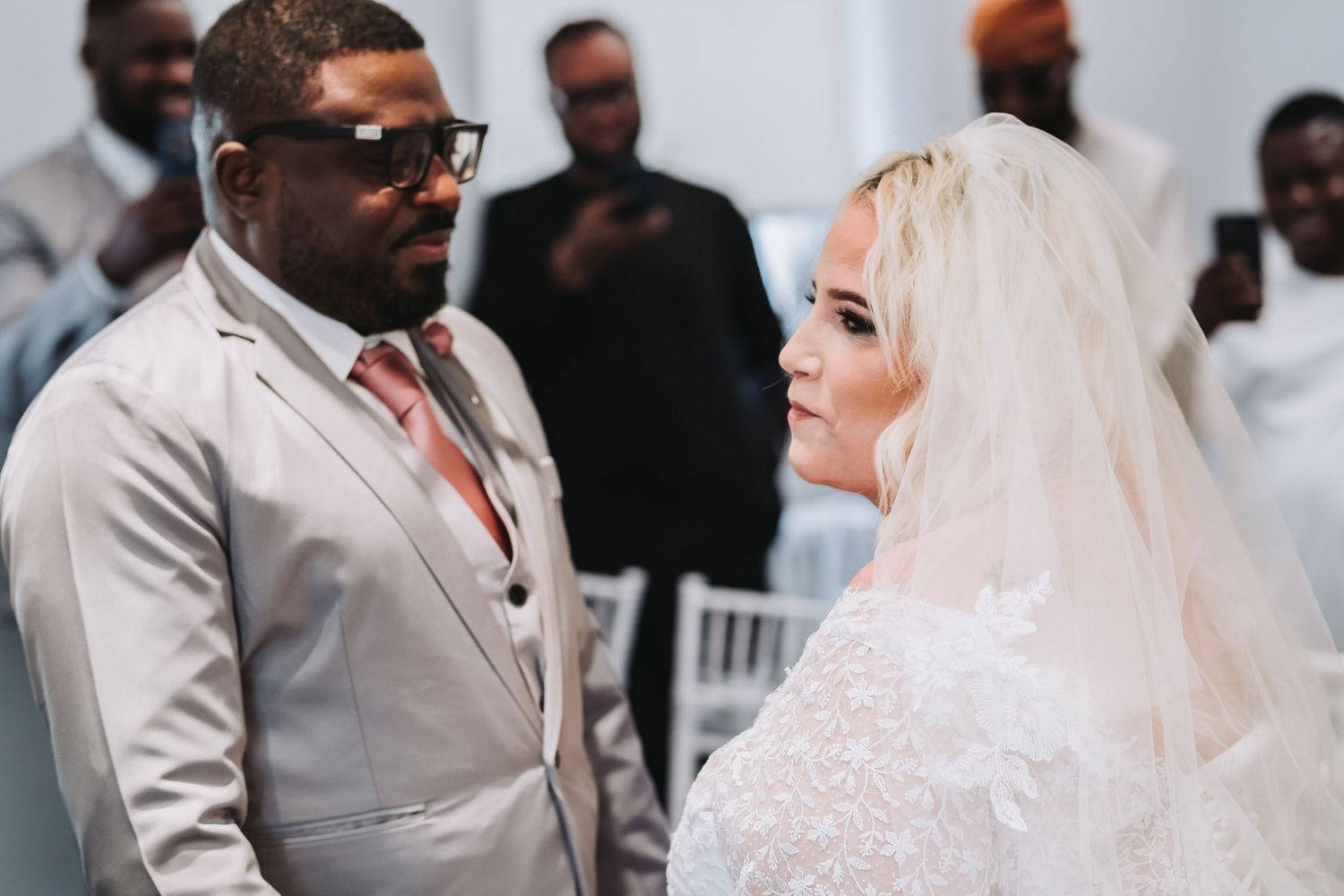 Wedding ceremony participants gather in formal attire with light gray suits and white decor.