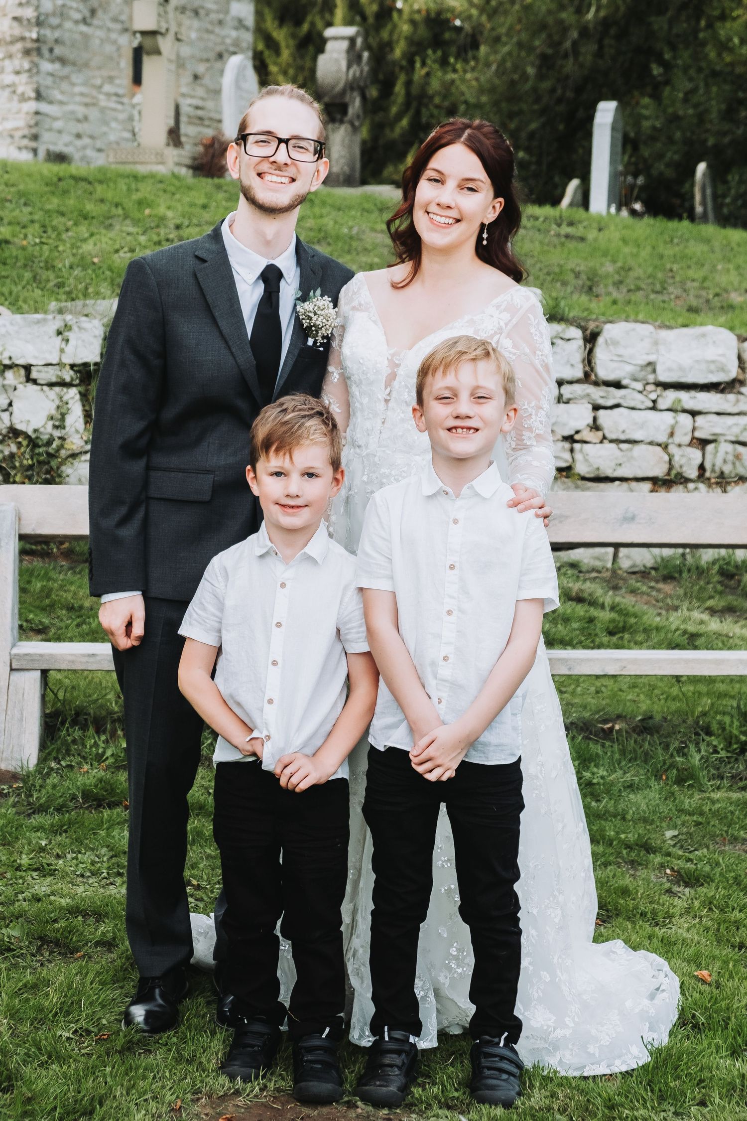 A family dressed in formal black and white attire poses together outdoors against a rustic stone wall backdrop.