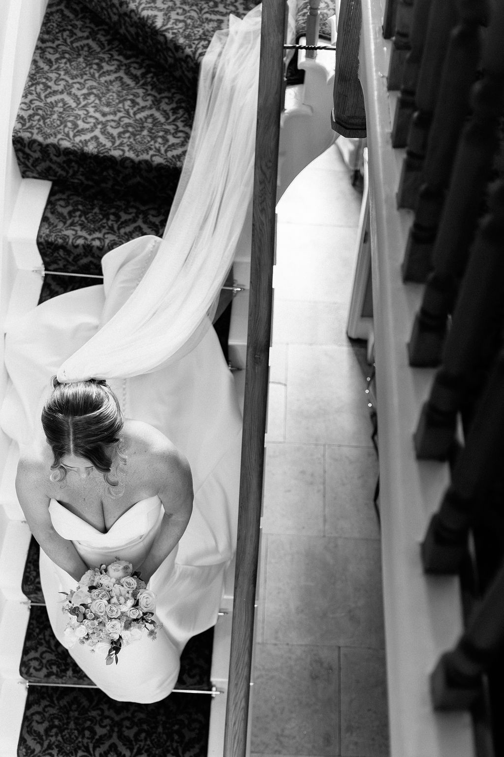 Black and white aerial view of a bride in a white gown holding a bouquet, descending an ornate staircase.