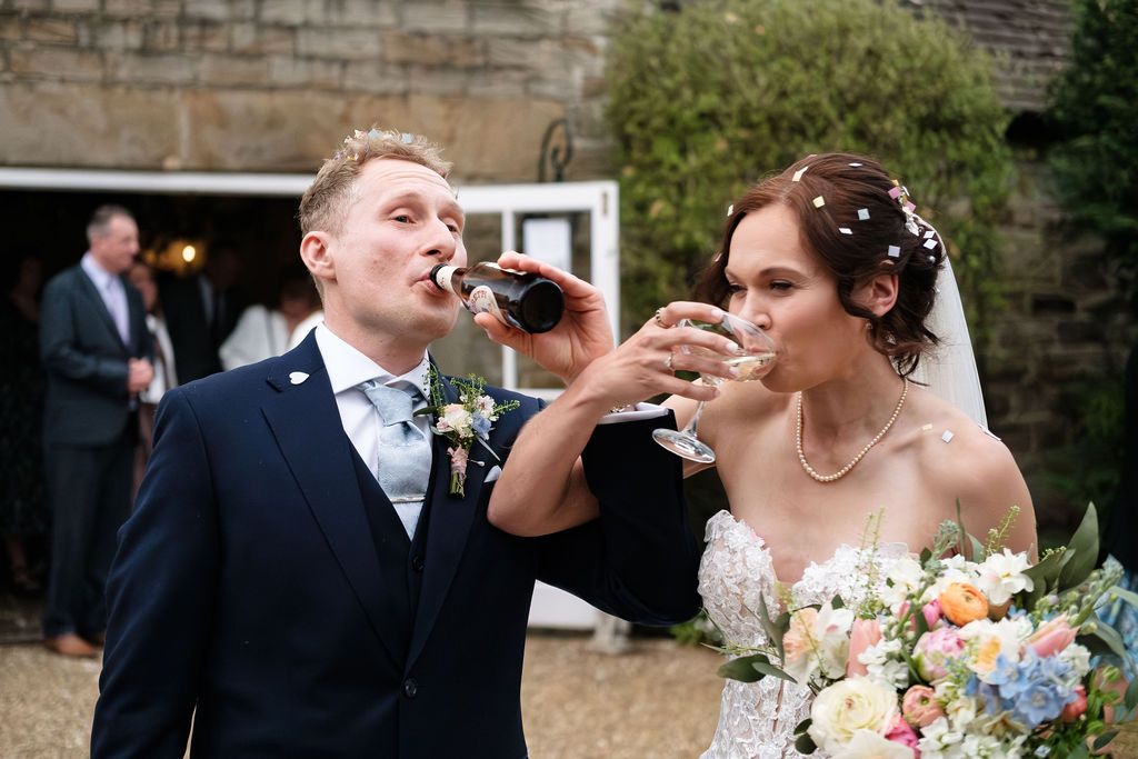 Bride and groom toasting with champagne glasses outdoors at their wedding venue, bride holding colorful bouquet.