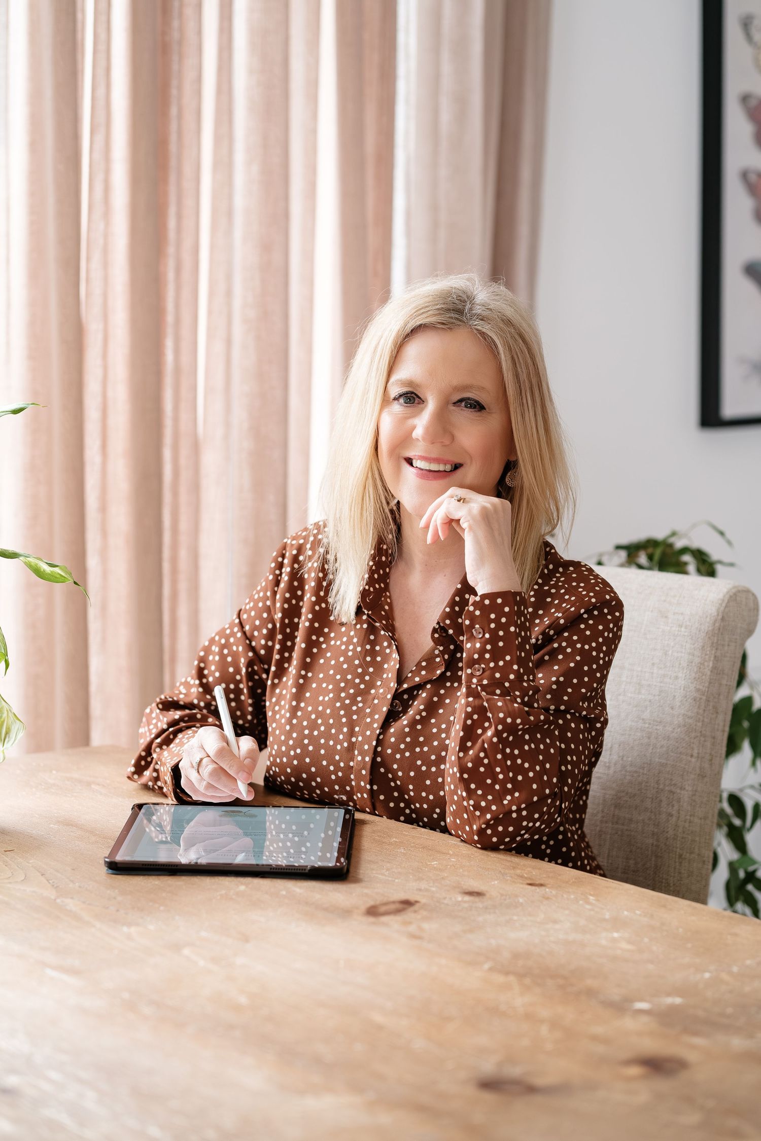 Smiling blonde woman in brown polka dot blouse using a digital tablet with stylus at a wooden desk.