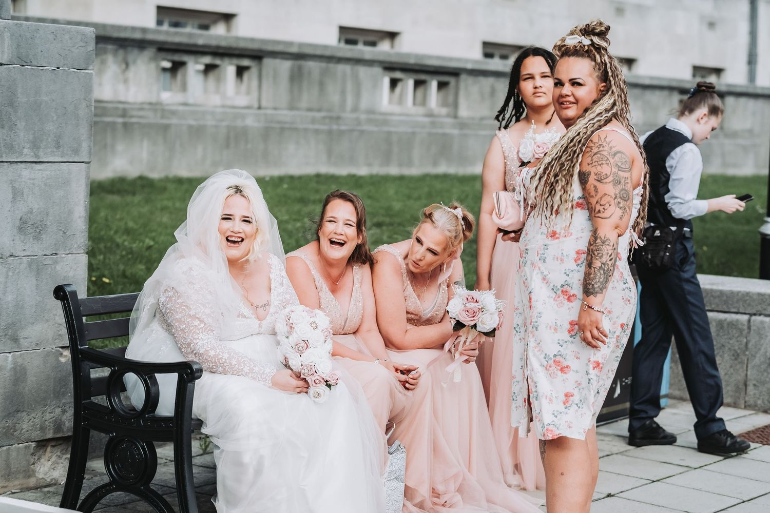 A group of bridesmaids in pink dresses laugh joyfully while sitting on a stone ledge with the bride in white.