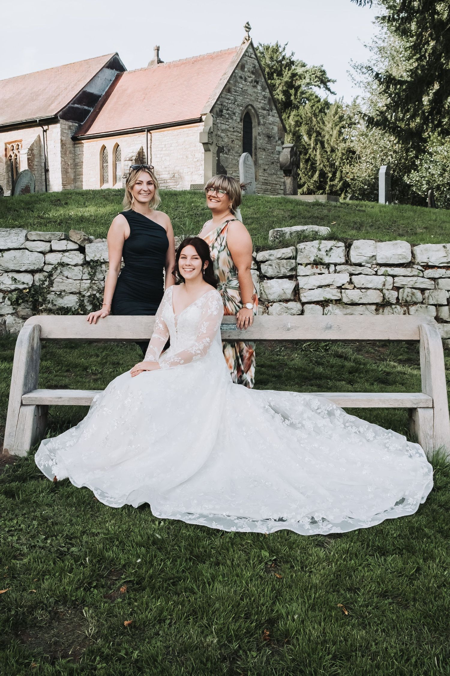 A beautiful wedding dress spreads across a rustic wooden bench against a stone wall backdrop.