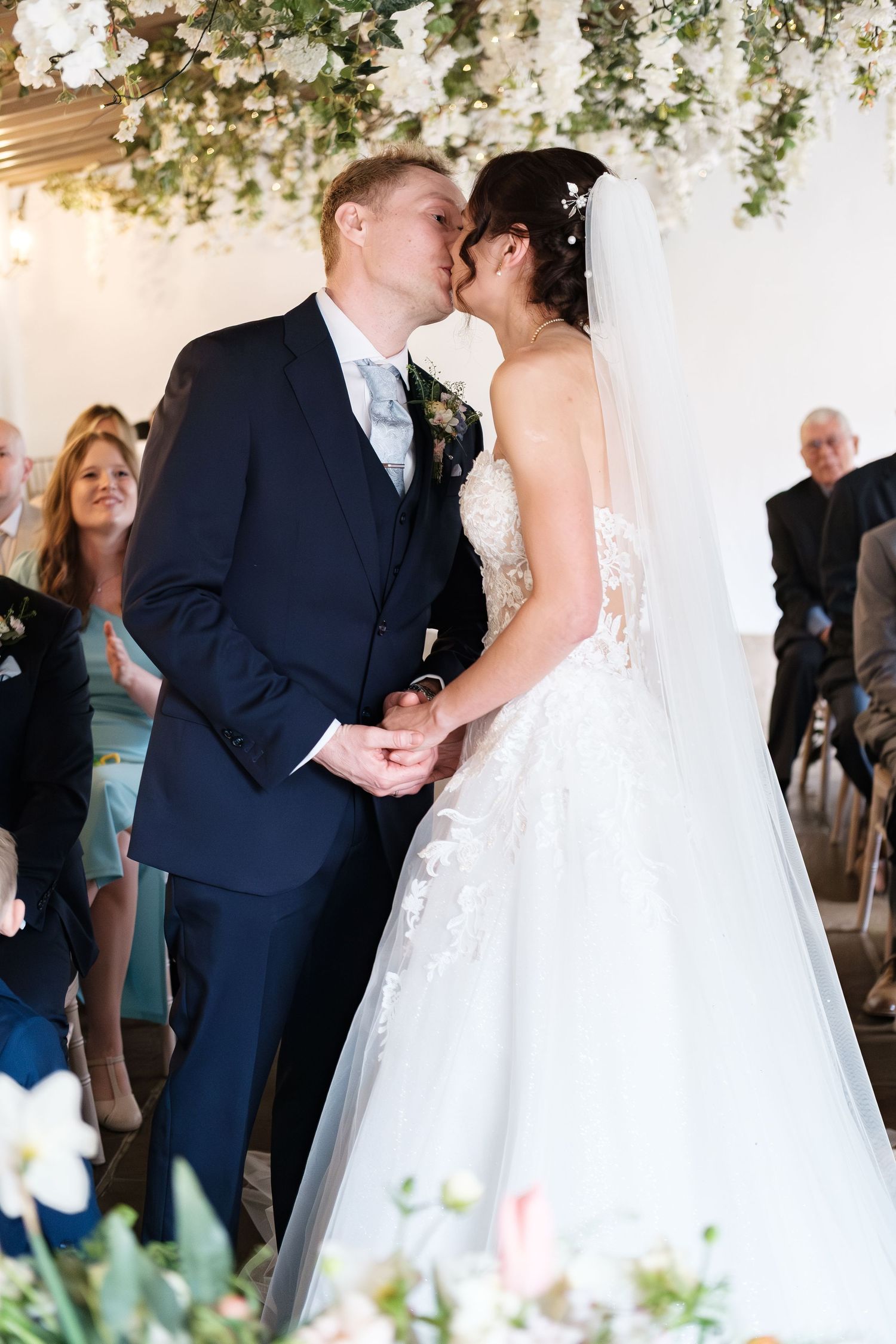 Bride and groom share first kiss at indoor wedding ceremony surrounded by floral arrangements and guests.
