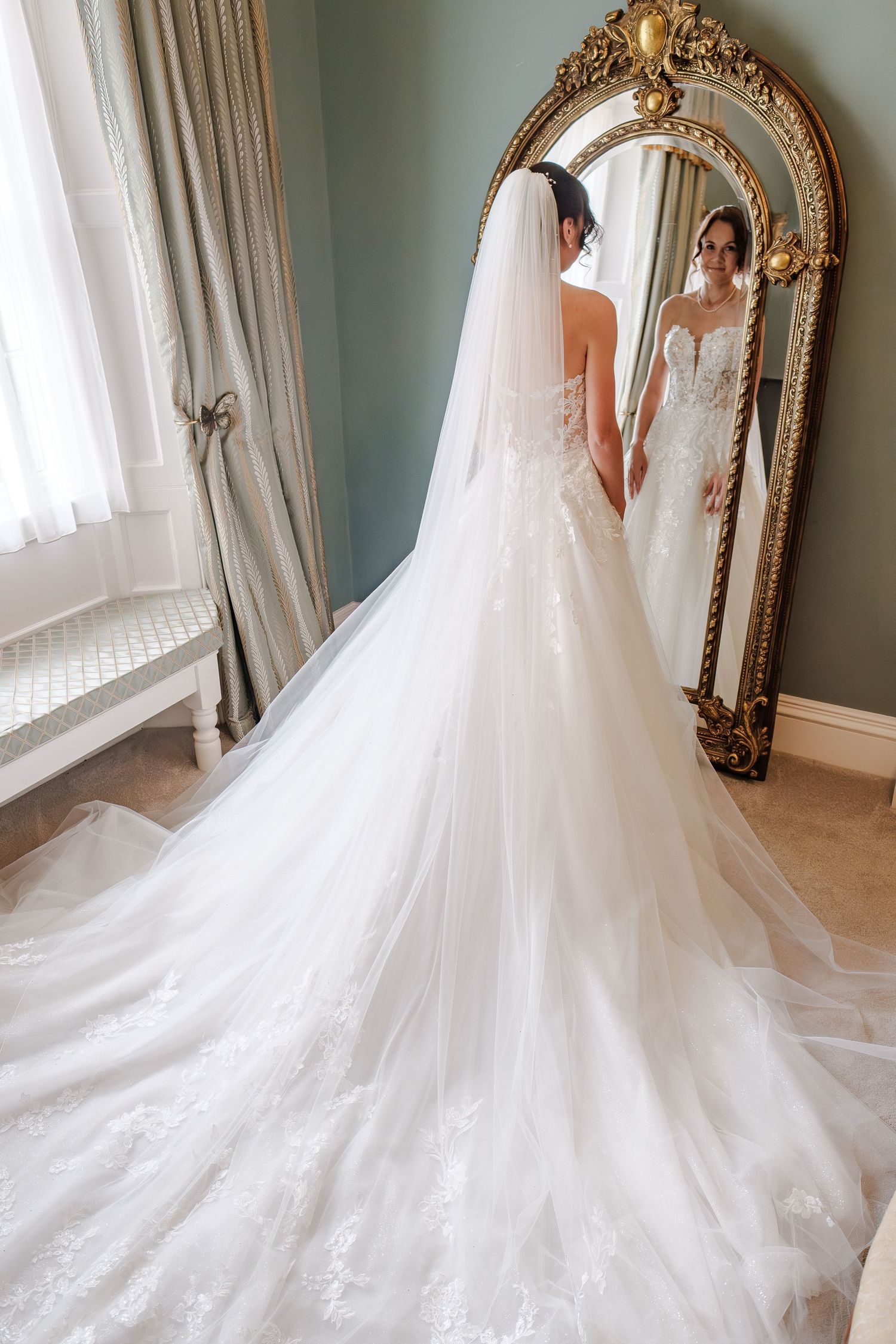 Bride in elegant lace wedding gown with long train and veil, viewed from behind in ornate gold mirror reflection.