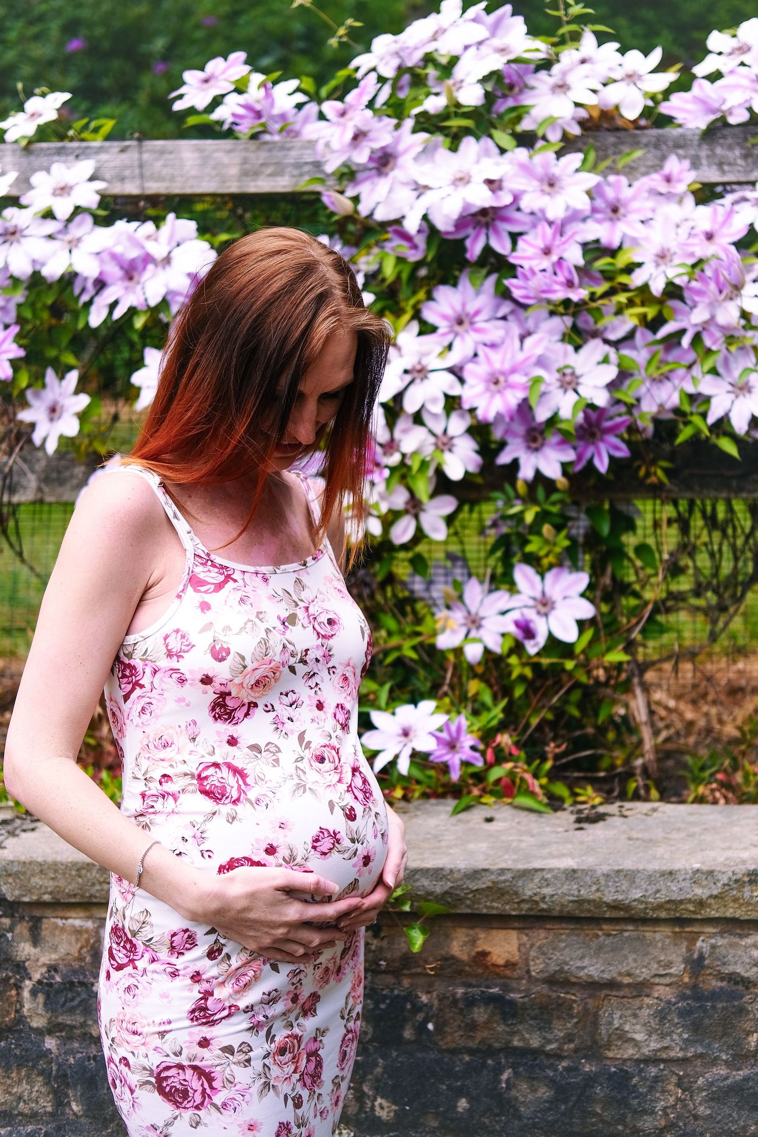 Maternity pose showcasing floral dress against blooming purple clematis backdrop.
