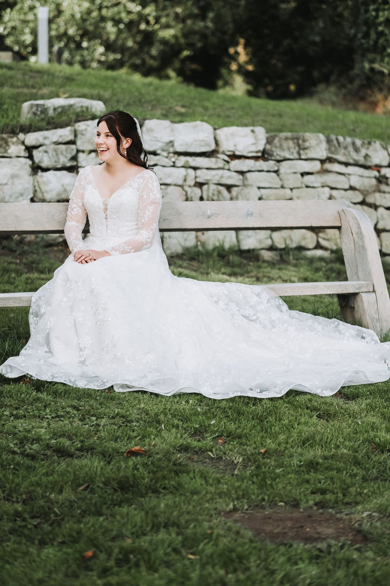 Elegant white lace wedding gown flowing over green grass near a stone wall.