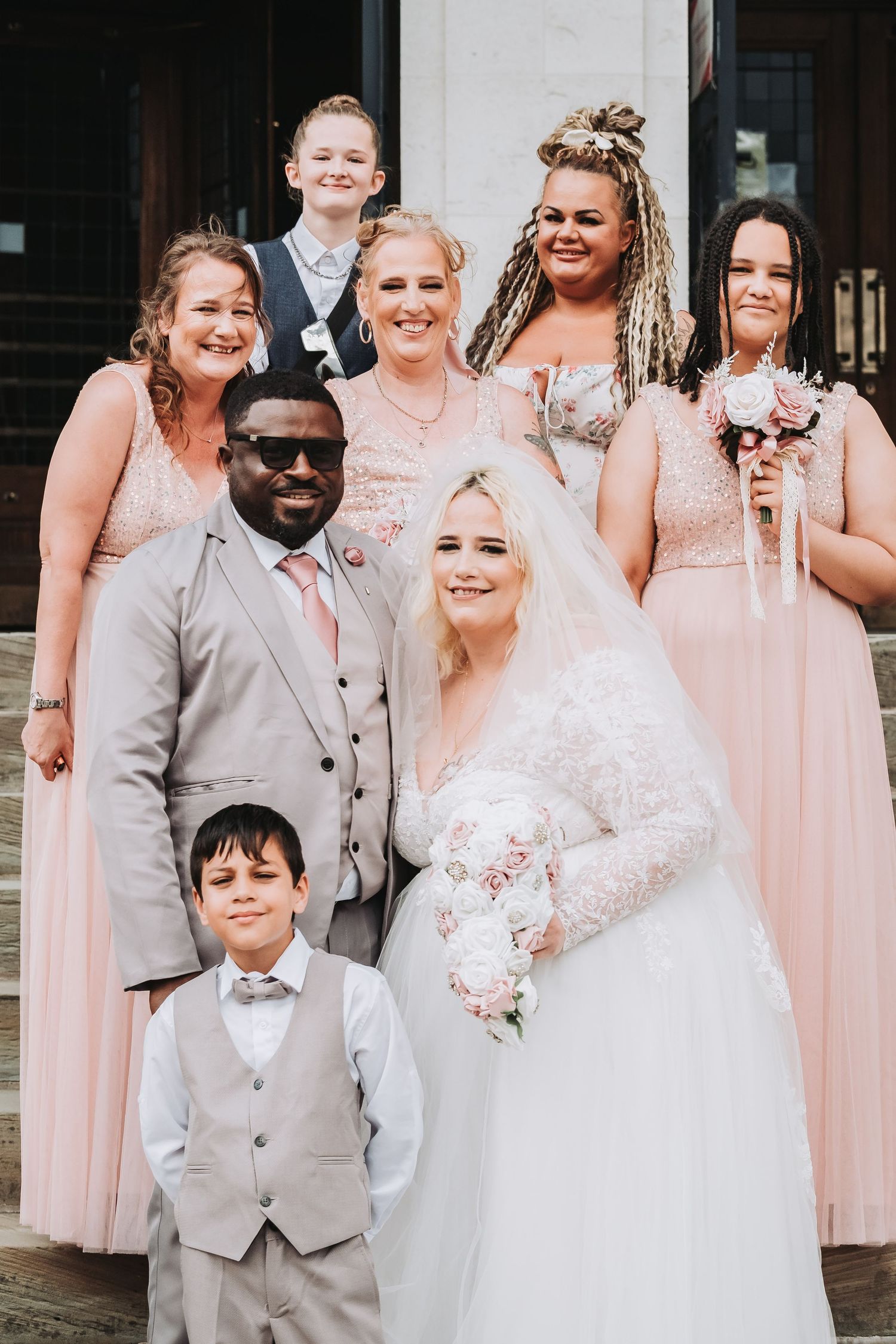 Wedding party poses together in pink bridesmaid dresses and gray suits outdoors.