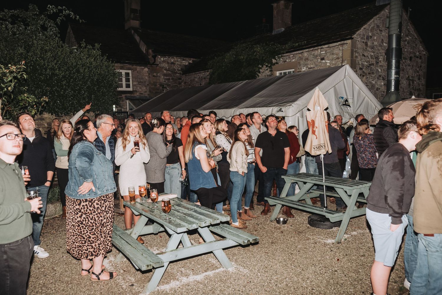 Outdoor nighttime party with picnic tables and guests socializing in casual attire.