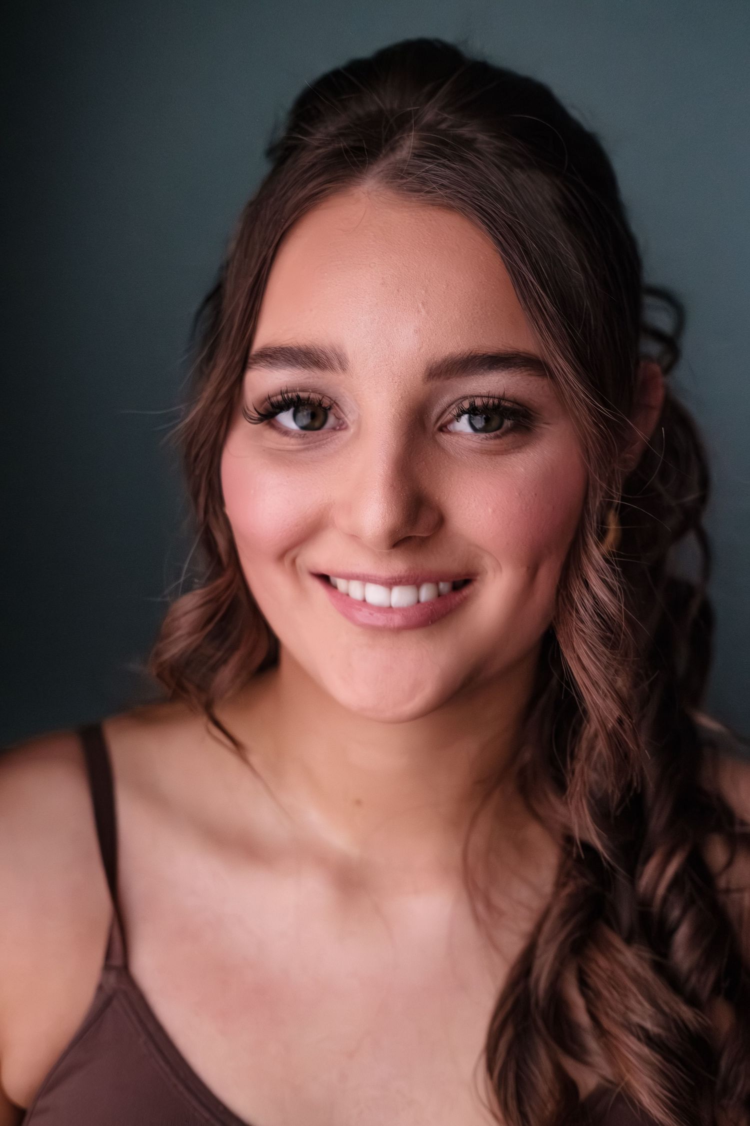 Young woman with wavy brown hair and green eyes smiling warmly against a dark teal background.